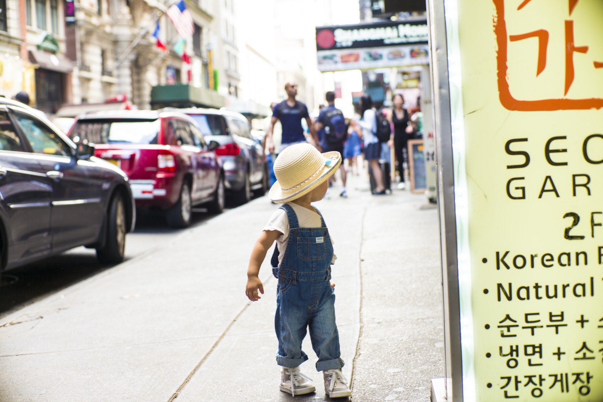 A toddler in overalls and a straw hat on a city street