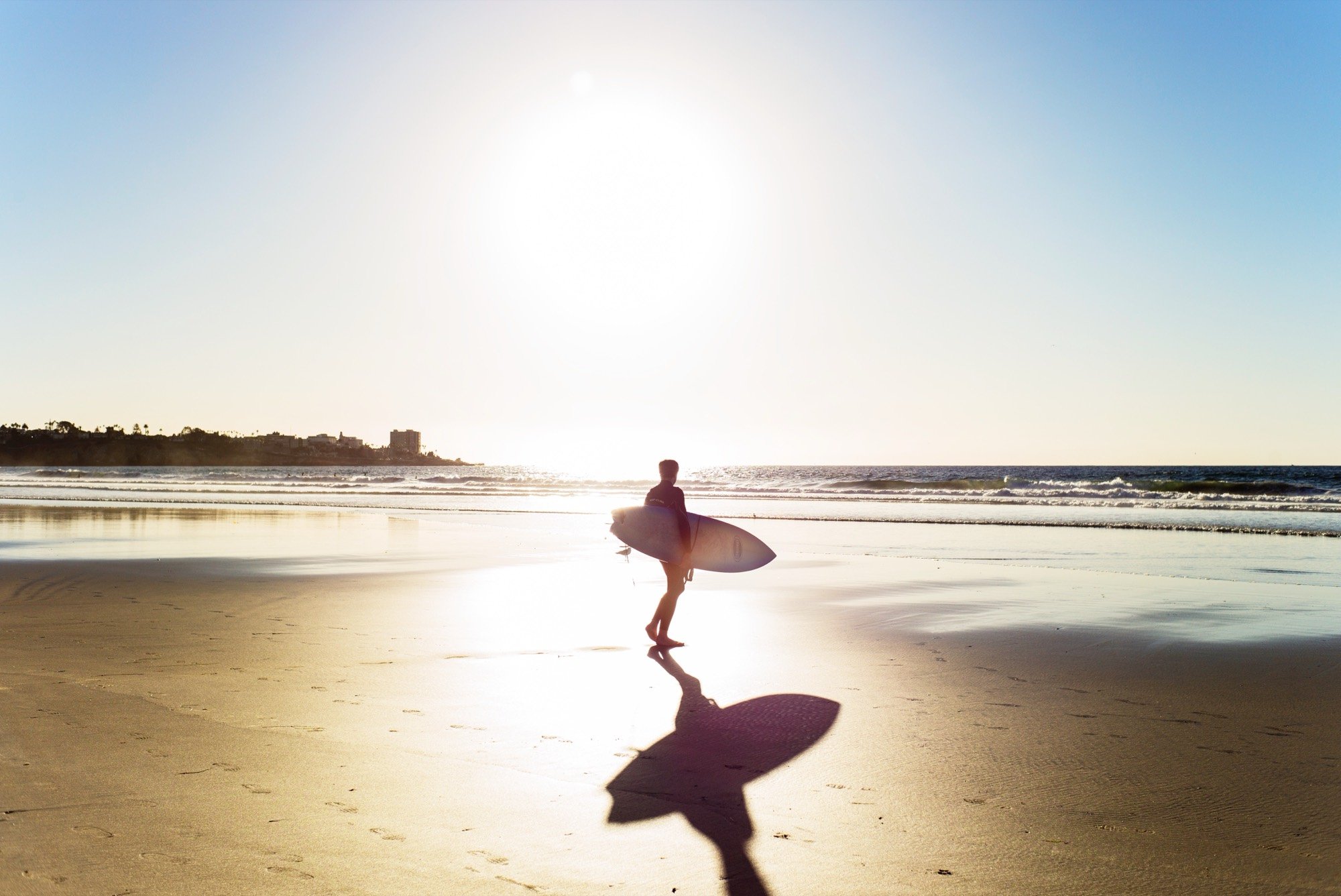 A surfer crossing a sunlit beach into the glare